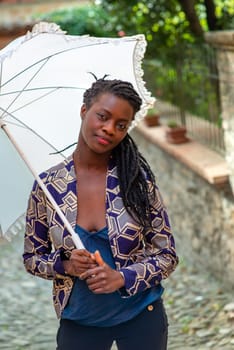 Young woman with dreadlocks braided hairstyle holding umbrella parasol. Happy young woman feeling confident in her style. Fashionable woman standing in the street of old village against stone wall.