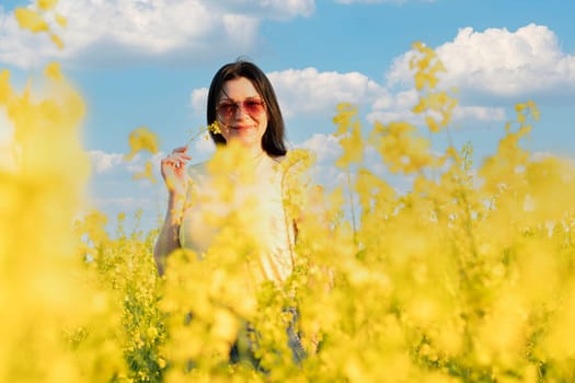 A beautiful girl in sunglasses stands in a field of yellow flowers against a blue sky, smiles happily and inhales the fresh spring and clean air. Peace concept. The color of the flag of Ukraine. No war.