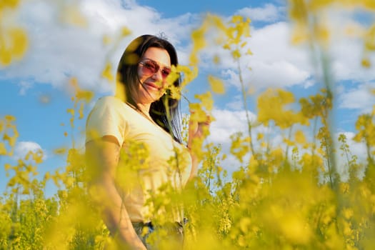 A beautiful girl in sunglasses stands in a field of yellow flowers against a blue sky, smiles happily and inhales the fresh spring and clean air. Peace concept. The color of the flag of Ukraine. No war.