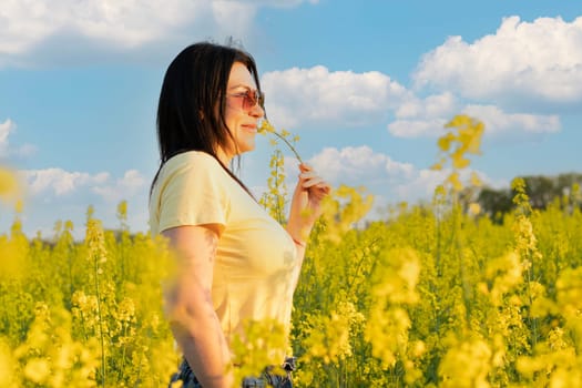 A beautiful girl in sunglasses stands in a field of yellow flowers against a blue sky, smiles happily and inhales the fresh spring and clean air. Peace concept. The color of the flag of Ukraine. No war.