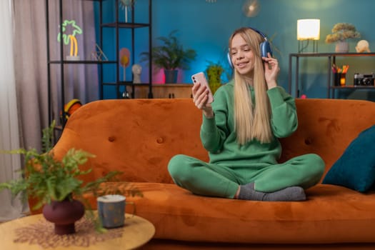 Happy overjoyed Caucasian woman in wireless headphones relaxing sitting on couch at home apartment choosing listening favorite energetic disco dancing music. People weekend daytime leisure activities