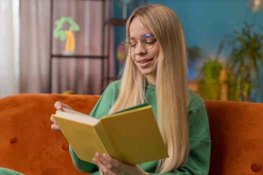 Young Caucasian woman relaxing interesting book turning pages smiling enjoying literature, taking a rest on comfortable couch. Portrait of peaceful cheerful girl at home apartment living room on couch
