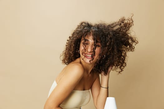 Woman dries curly hair with blow dryer, home beats styling products, smile on beige background. High quality photo