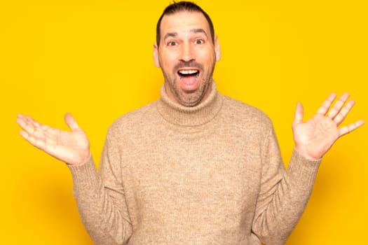 Funny smiling man in beige sweater showing palms up, unexpected surprise gesture isolated over yellow background. Attractive happy man listening to good news, catching something