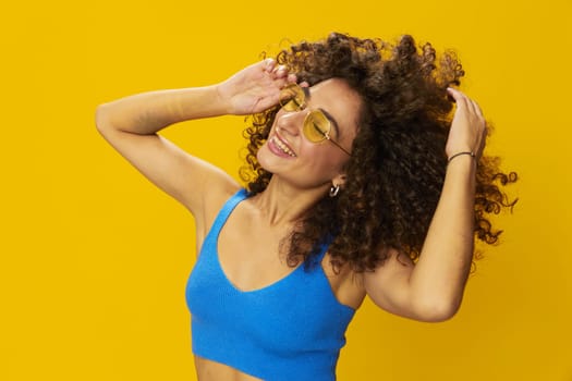 Woman with curly afro hair in a blue T-shirt on a yellow background dancing flying hair with sunglasses yellow, hand signs, look into the camera, smile with teeth and happiness, copy space. High quality photo