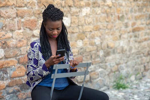 Close up portrait of beautiful young african american woman reading answering text message on mobile phone app over a wall outdoors