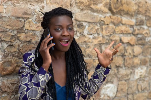 confident African young adult woman holding cell phone calling , receives good news, smiling, happy, outdoors in the village over a wall.