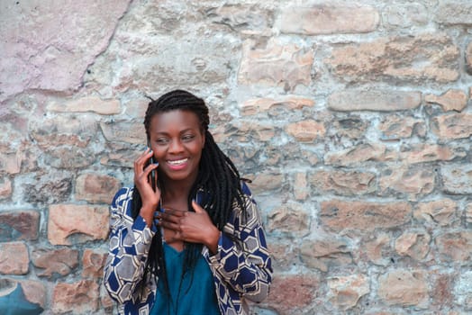 confident African young adult woman holding cell phone calling , receives good news, smiling, happy, outdoors in the village over a wall.