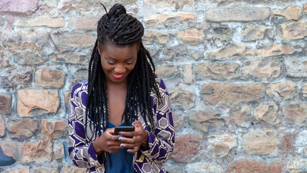 Close up portrait of beautiful young african american woman reading answering text message on mobile phone app over a wall outdoors