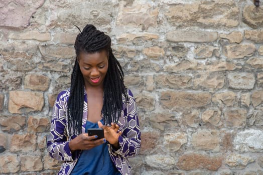 Close up portrait of beautiful young african american woman reading answering text message on mobile phone app over a wall outdoors