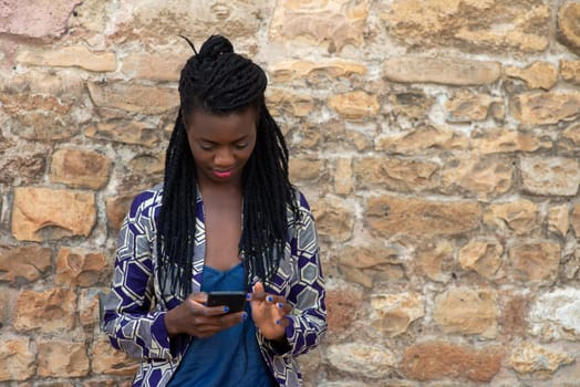 Close up portrait of beautiful young african american woman reading answering text message on mobile phone app over a wall outdoors