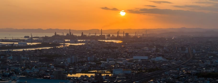 Panoramic aerial view of low sun in orange sky over industrial smokestacks and city at sunset. High quality photo