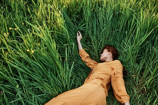 a sweet, calm woman in an orange dress lies in a green field with her arms outstretched and her eyes closed, enjoying the silence and peace. Horizontal photo taken from above. High quality photo