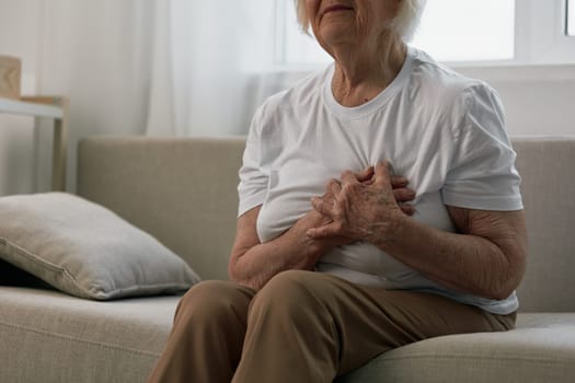 Elderly woman severe chest pain sitting on the sofa, health problems in old age, poor quality of life. Grandmother with gray hair holds her chest with her hands, women's health, breast cancer. High quality photo