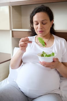 Multi ethnic adult pregnant woman in third trimester of her pregnancy, sitting on the bed at home, eating healthy vegetable salad, enjoying healthy lifestyle and maternity leave. Expecting a baby