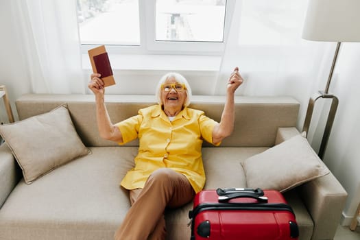 Happy senior woman with passport and travel ticket packed a red suitcase, vacation and health care. Smiling old woman joyfully sitting on the sofa before the trip raised her hands up in joy. High quality photo