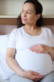 Close-up portrait of beautiful ethnic woman looking away, caressing her belly, enjoying happy moments of pregnancy and anticipation to be a mother. Motherhood. Childbirth. Expectation of childbirth