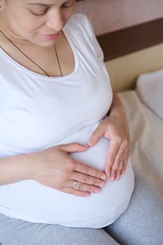 Top view. Charming pregnant woman in white t-shirt, shows a heart made from her fingers over her belly, feeling connection with her future baby, expressing positive emotions of pregnancy and maternity