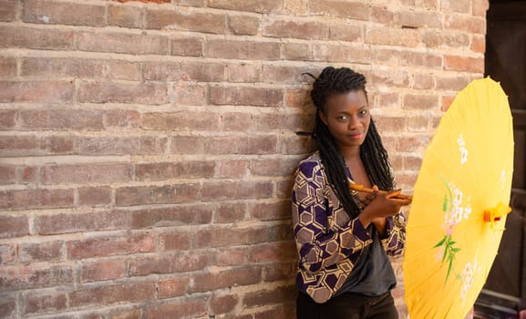 Young woman with dreadlocks braided hairstyle holding umbrella parasol. Happy young woman feeling confident in her style. Fashionable woman standing in the street of old village against stone wall.