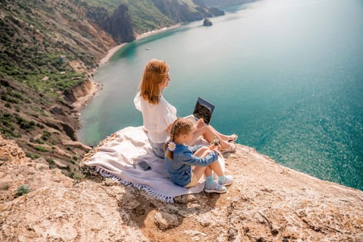 Freelance woman with her daughter working on a laptop by the sea, typing on the keyboard, enjoying the beautiful view, highlighting the idea of remote work