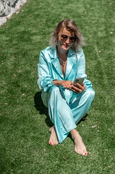 Woman grass. Happy woman with blond curly hair in glasses and in a blue overalls sits on the summer fresh green grass with a phone in her hands