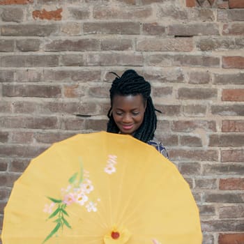 Young woman with dreadlocks braided hairstyle holding umbrella parasol. Happy young woman feeling confident in her style. Fashionable woman standing in the street of old village against stone wall.