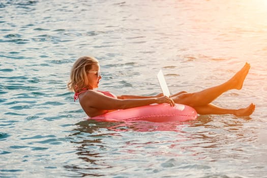 Woman works on laptop in sea. Freelancer, young blond woman in sunglases floating on an inflatable big pink donut with a laptop in the sea at sunset. Freelance, travel and holidays concept