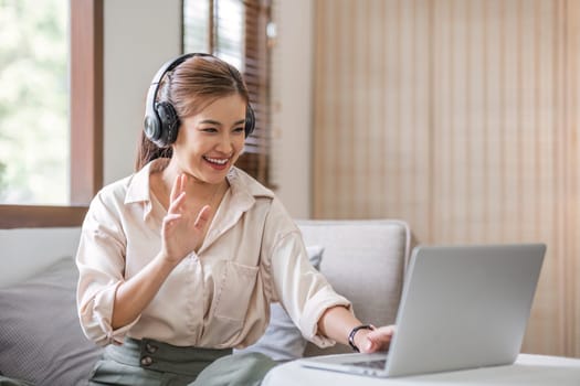 Smiling young asian woman using laptop web camera while wearing headphones and sitting on the rug beside to the sofa at homes..