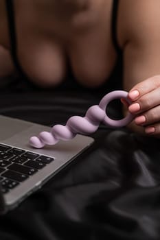 Woman holding lilac anal beads next to laptop while lying on black sheet