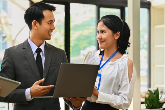 Asian male manager holding laptop speaking, sharing ideas with colleagues at corporate meeting.