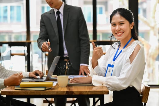 Smiling asian woman employee showing thumb up while working on laptop and her colleagues standing in background.