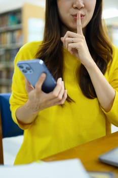 Young woman holding smartphone and making shush gesture as sign of silence, secrecy, secret, keep quiet sign.