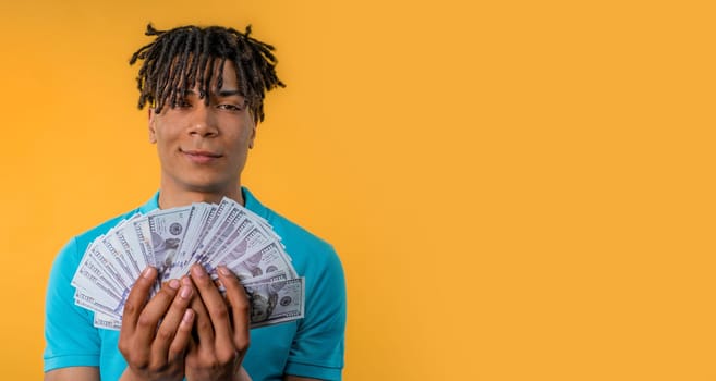 Satisfied man with USD currency. Young millenial african american guy or student holding money - dollars banknotes on yellow wall. Symbol of success, gain, salary, benefit, investments. Copy space