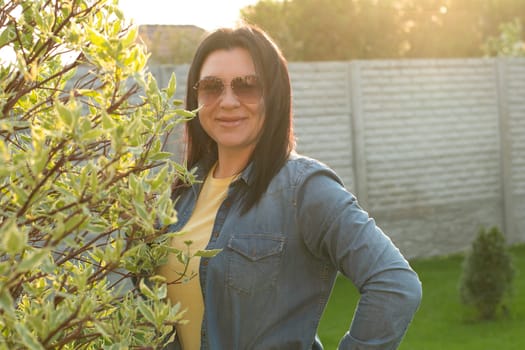 Camping. A beautiful girl in jeans and sunglasses, stands near a blooming green tree, smiles happily. Fresh air concept. Sunset.