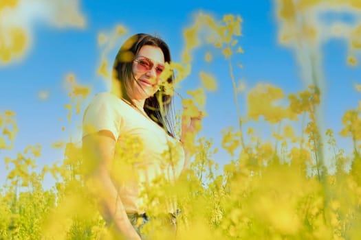 A beautiful girl in sunglasses stands in a field of yellow flowers against a blue sky, smiles happily and inhales the fresh spring and clean air. Peace concept. The color of the flag of Ukraine. No war.