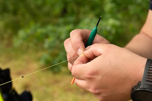 Fishing concept. Close-up male hands knit a float with a fishing line to a fishing rod against the background of green trees. Bait for fish. Soft focus. copy space.