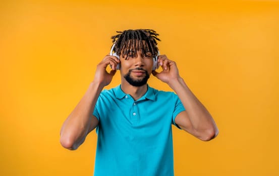 Positive bearded african american man listening music, enjoying with headphones on yellow studio background. Radio, wireless modern sound technology, online player. High quality