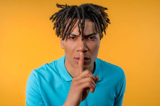 Handsome african american teenager guy with gesture of shhh, secret, silence, conspiracy, gossip concept. Smiling guy holding finger on lips, yellow studio background. High quality
