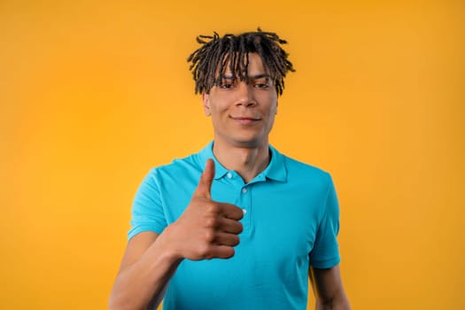 Positive african american man makes hand sign like, thumbs up gesture. Happy student guy in blue, correct perfect choice, great deal, yellow studio background. High quality