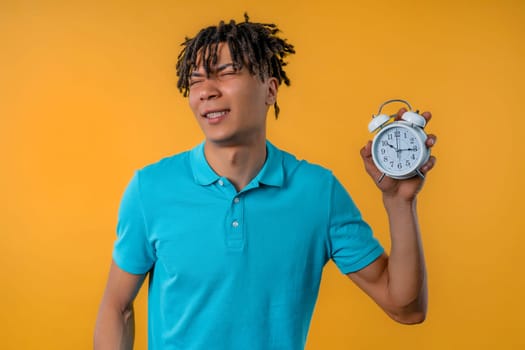 Woken up by alarm clock sleepy african man holding it in hand. Yellow background. 10 o'clock in morning. Lazy guy didn't get enough sleep, concept of passing time. High quality