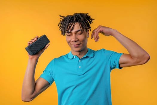 African american man dancing, enjoying on yellow studio background. Guy moves to rhythm of music. Young teenager listening to music by wireless portable speaker - modern sound system. High quality