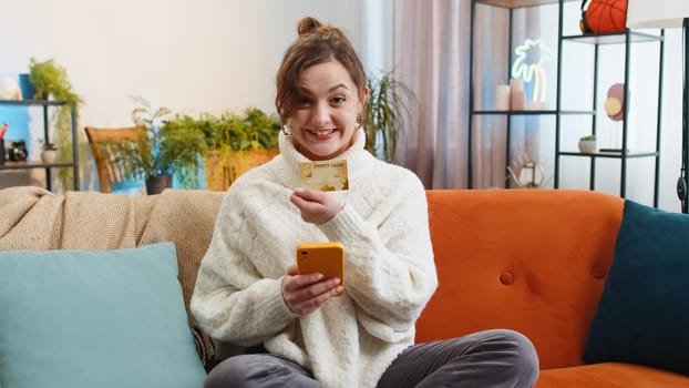 Woman using credit bank card and smartphone while transferring money, purchases online shopping cashless, order food delivery at home apartment indoors. Happy young girl sitting in room on couch