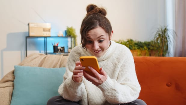 Happy woman use mobile smartphone typing browsing say Wow yes found out great big win good news celebrate lottery jackpot doing winner gesture. Portrait of young girl at home in room sitting on sofa