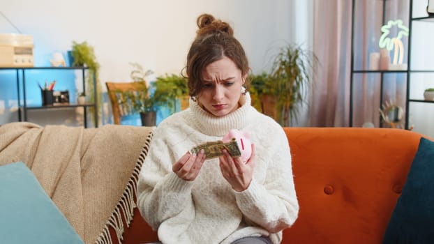 Poor young woman insufficient amount of money, holding piggybank and one dollar banknote at home apartment. Financial crisis. Bankruptcy. Poverty and destitution. Girl in living room sitting on sofa