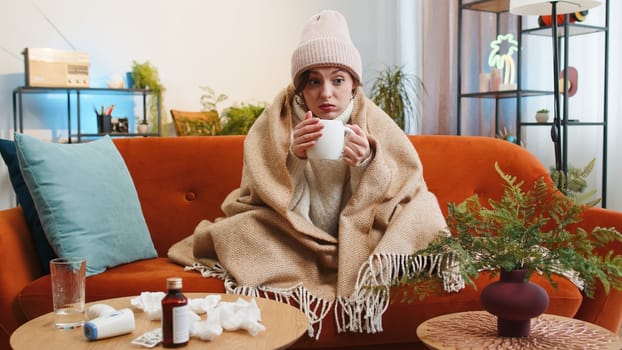 Young ill woman suffering from cold or allergy sitting on sofa in living room at home drinking hot tea. Sick brunette girl wear hat wrapped in plaid blows sneezes wipes snot into napkin. Quarantine