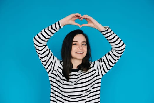 Joyful Woman in Striped Sweater Spreading Love - Smiling Girl with Raised Arms Creating Heart Shape, Radiating Happiness and Positivity - Isolated Blue Background. Perfect for Positive Vibes Concepts