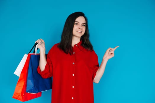 Cute Woman in Red Shirt with Shopping Bag - Pointing to Empty Space - Perfect for Promoting Sales and Discounts - Isolated on Blue Background. Smiling Woman with Shopping Bag