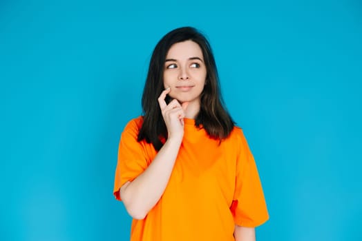 Attractive Young Woman in Trendy Orange Clothes, Pensive and Skeptical Expression, Empty Space for Copy, Isolated on Blue Background - Portrait Photo.