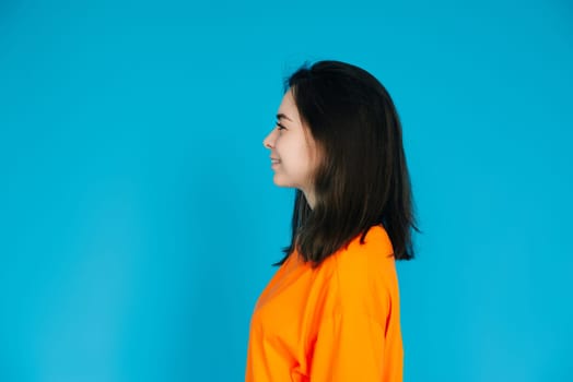 Radiant Young Woman in Stylish Orange Attire Smiling with a Beaming Toothed Smile, Profile Portrait, Isolated on Blue Background.