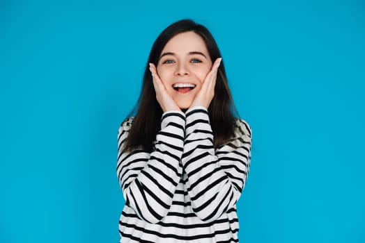 Expressive Portrait of a Surprised and Joyful Young Woman with Palms on Cheekbones, Isolated on Blue Background.